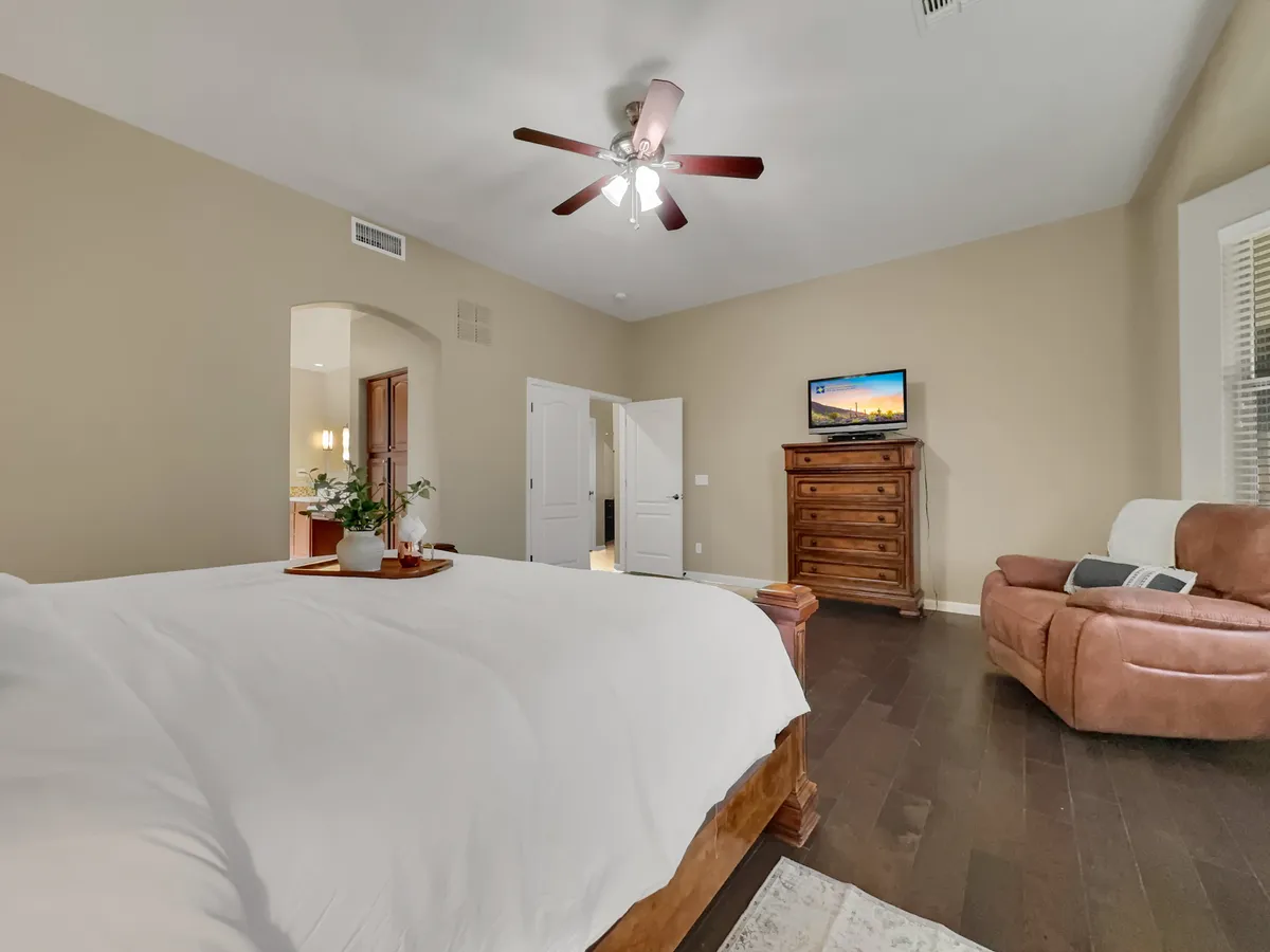 A serene king-sized bedroom featuring white linens, a wooden dresser, a cozy recliner, and a wall-mounted TV. The space receives gentle natural light, creating an inviting retreat perfect for unwinding after a day of activities.