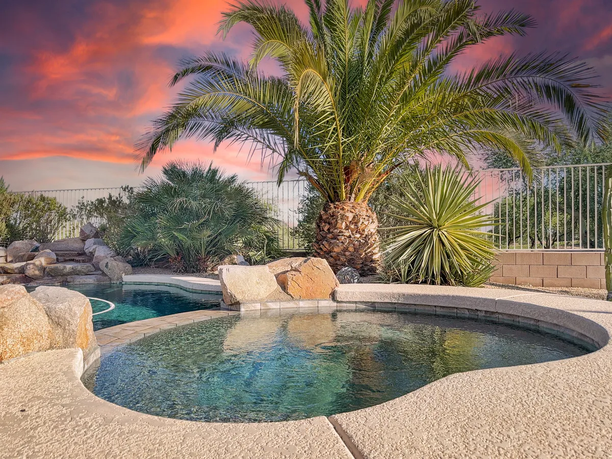 A luxurious hot tub sits adjacent to a spacious swimming pool, all framed by tropical palm trees and rugged rock features. The view suggests warmth and relaxation, with ample space for guests to unwind and enjoy both water amenities together in a scenic setting.