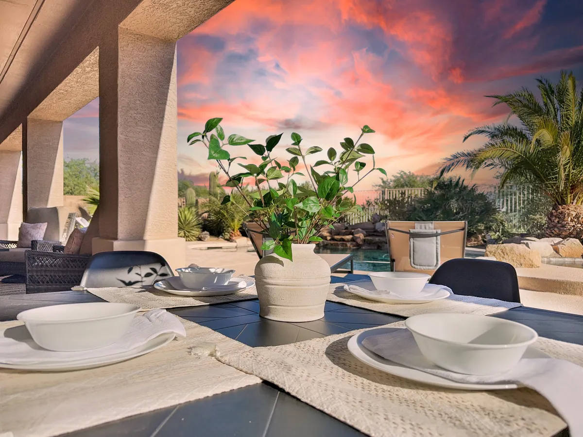 A serene exterior dining area featuring a table elegantly set with white bowls and a plant centerpiece, positioned under a vibrant sunset sky. Lush greenery surrounds the space, and the poolside location enhances the ambiance for outdoor meals and gatherings.