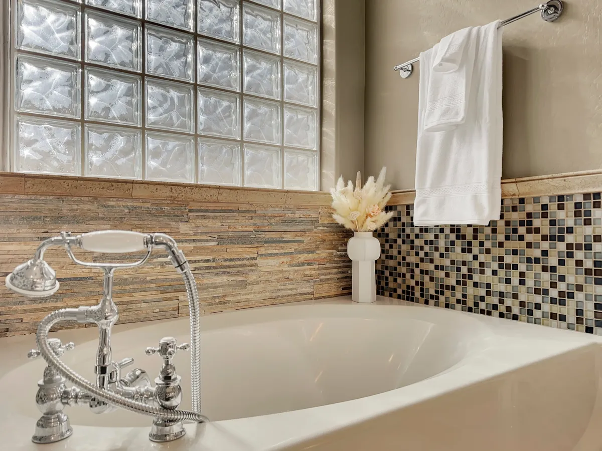 A modern bathroom featuring a freestanding bathtub, decorative tile backsplash, a glass block window for gentle natural light, a vase with fresh flowers, and neatly arranged towels. The space exudes spa-like tranquility.