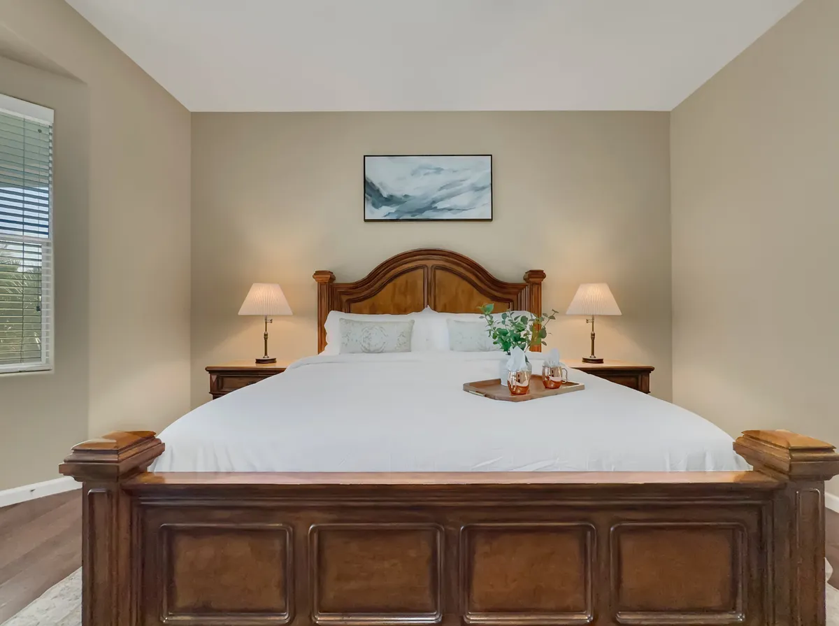 A queen bedroom with a wooden bed frame, decorative painting, soft lamp lighting, and serene bedding, all bathed in natural light filtering in through the window. The décor is understated and soothing, evoking a sense of peace.