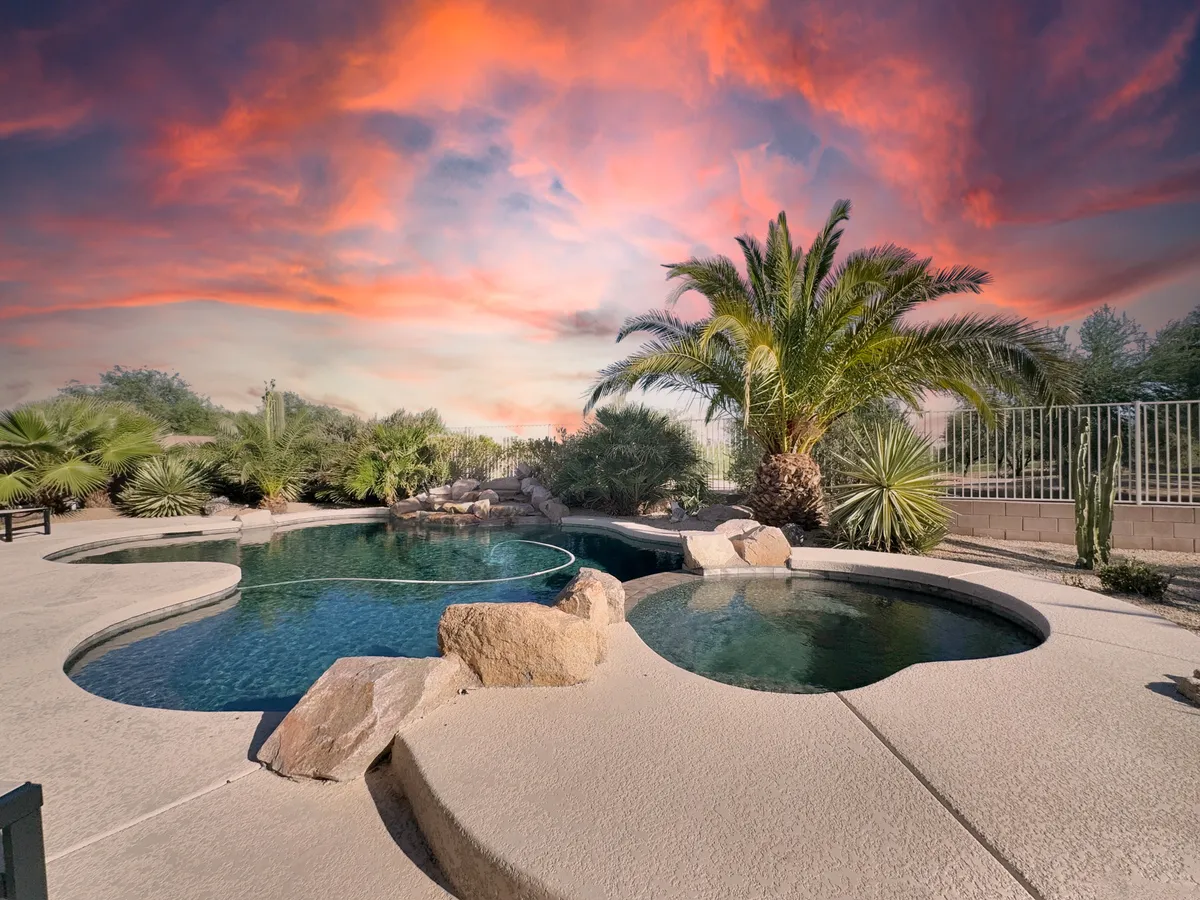 This tranquil exterior shows a shimmering pool surrounded by palm trees and distinctive rock formations, with a beautiful sunset sky in the background. The setting is serene and lush, providing an idyllic space for swimming, sunbathing, or enjoying evening gatherings as day transitions into night.
