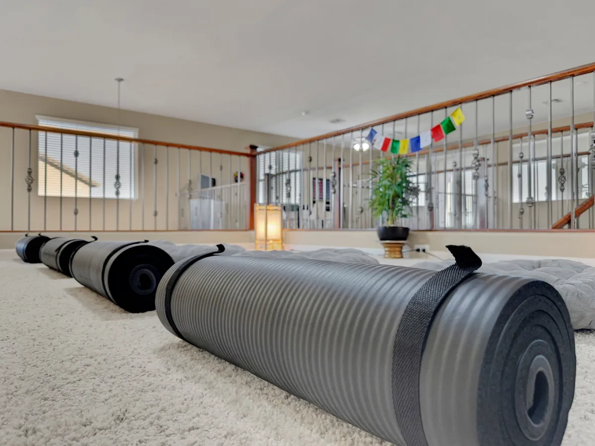 A sunlit, tranquil interior space set up for yoga or meditation with yoga mats arranged on the floor, decorative flags, a floor lamp, and an indoor plant. The ambiance is calm and inviting, perfectly suited for group or solo wellness sessions.