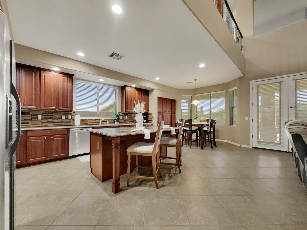 A bright, welcoming kitchen and dining area with a large island counter, wood cabinetry, and a dining table positioned near expansive windows. The space offers ample prep surfaces and natural light, providing an ideal backdrop for both cooking and sharing meals.