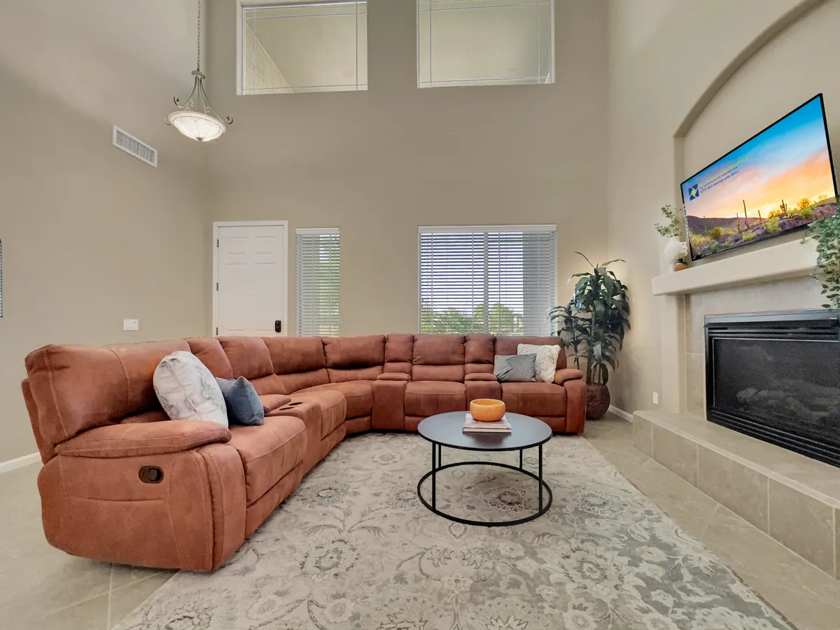 A comfortable living room with a spacious sectional sofa, a central coffee table, a fireplace, and large windows that allow natural light to stream in. The room is set up for relaxation, socializing, or watching television, with a serene view visible through the windows.